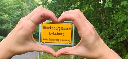Frauenhände, als Herz gehalten, die das Ortsschild von Glücksburg umrahmen mit grünem Wald im Hintergrund