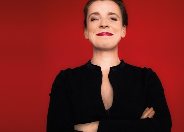 Halbporträt Comedienne Tina Teubner in schwarzer Bluse mit verschränkten Armen und Grinsen im Gesicht vor roter Wand 