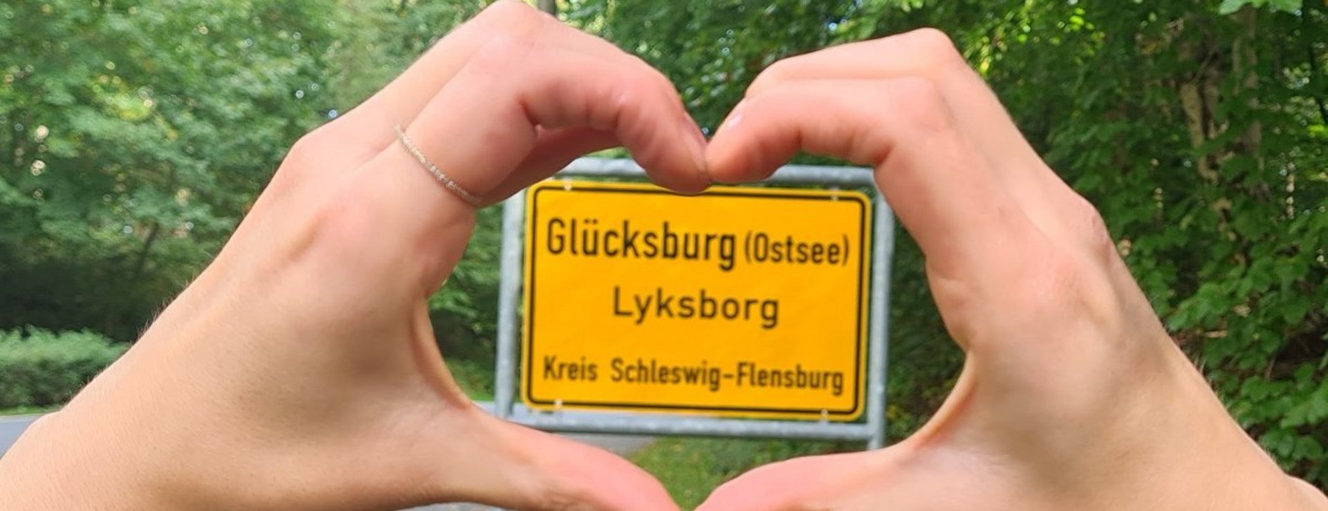 Frauenhände, als Herz gehalten, die das Ortsschild von Glücksburg umrahmen mit grünem Wald im Hintergrund